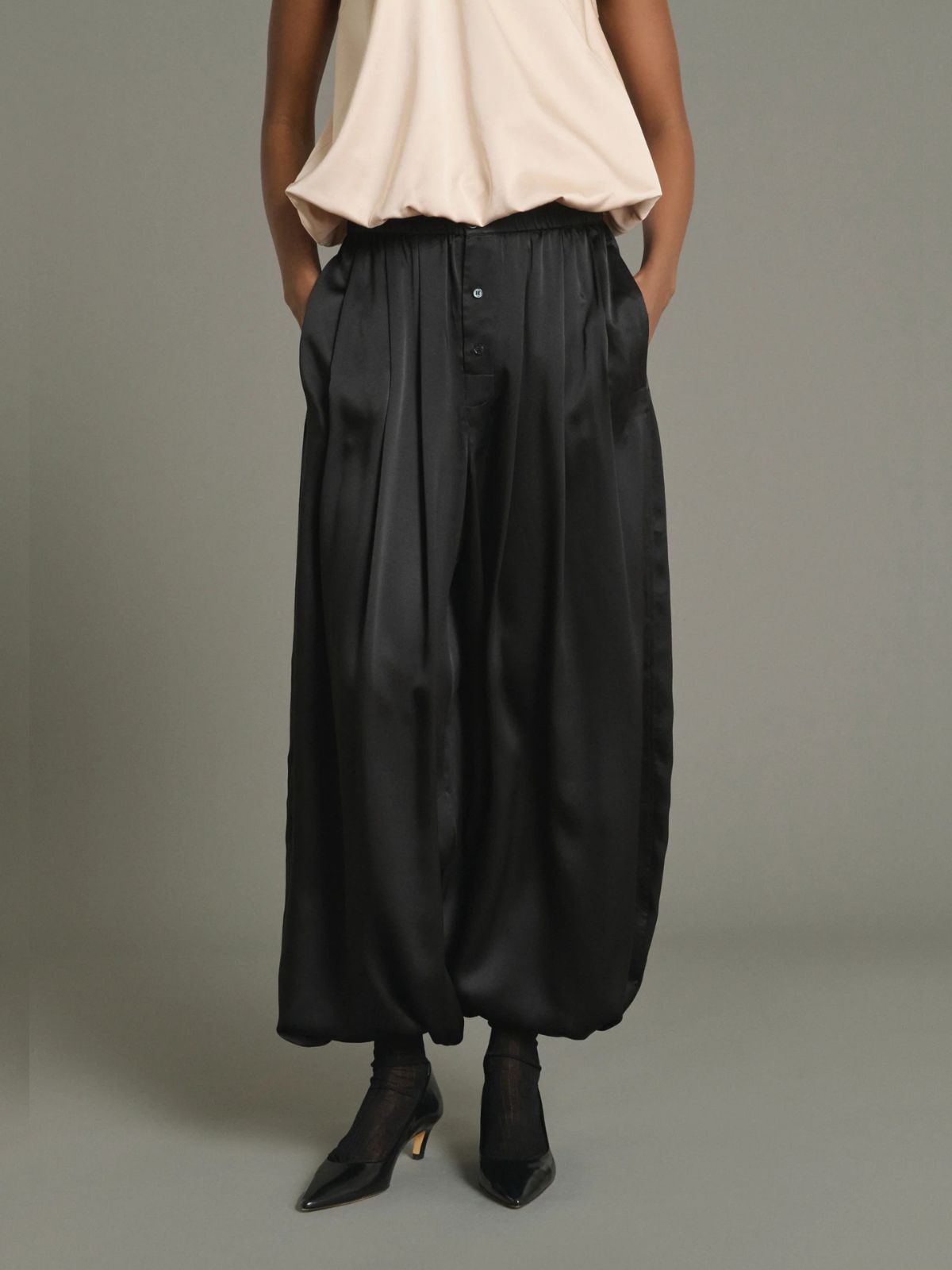 A model is shown from the waist down wearing black wide-leg satin pants with a button-up front, paired with a light beige top and black pointed-toe heels, against a solid gray studio background.