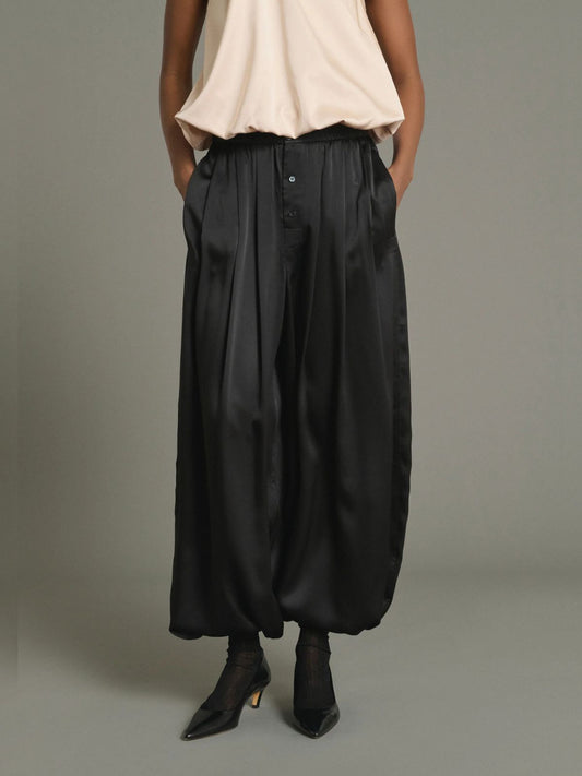 A model is shown from the waist down wearing black wide-leg satin pants with a button-up front, paired with a light beige top and black pointed-toe heels, against a solid gray studio background.